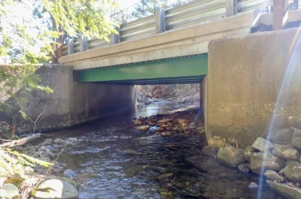 A concrete and metal bridge spans over a shallow creek with rocks and sunlight visible through nearby trees.