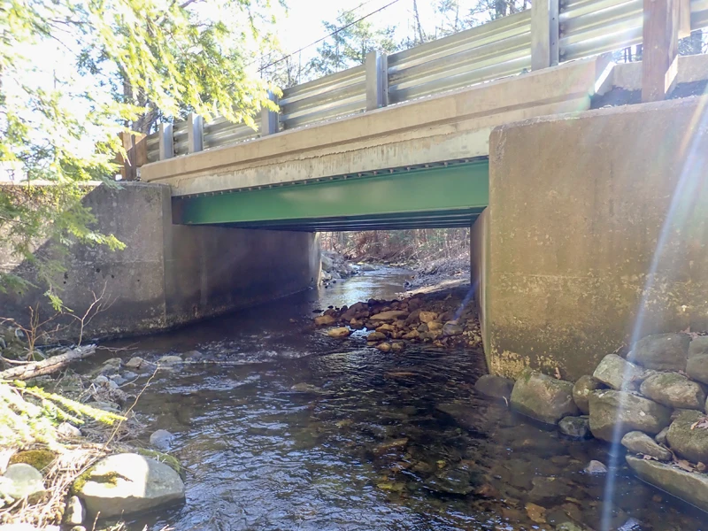 A concrete and metal bridge spans over a shallow creek with rocks and sunlight visible through nearby trees.