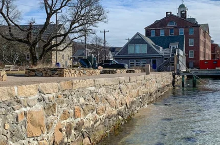 A stone seawall lines the edge of a waterfront with buildings, leafless trees, and parked cars in the background under a partly cloudy sky.