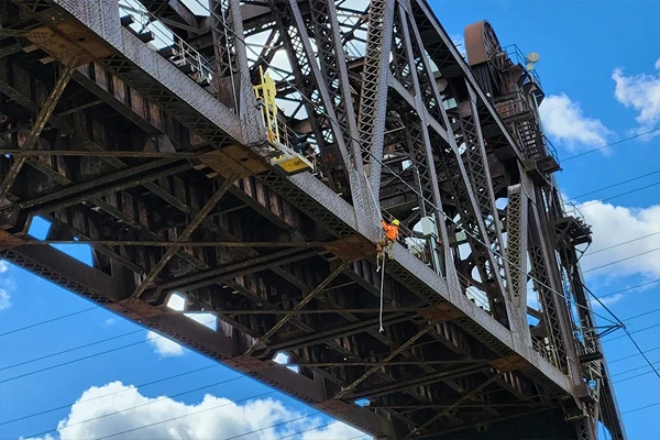 A worker in an orange vest performs maintenance on a tall steel bridge against a blue sky with clouds.