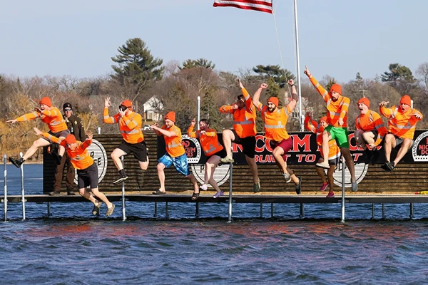 A group of people in orange shirts and hats jump off a dock into a lake, with an American flag and trees visible in the background.
