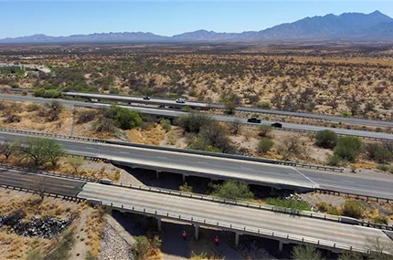 A highway overpass crosses above another road in a desert landscape, with sparse vegetation and distant mountains under a clear blue sky.