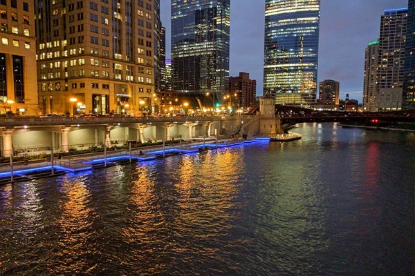 City buildings and riverwalk with blue lights reflected in the water at dusk, surrounded by illuminated skyscrapers and a bridge in the background.