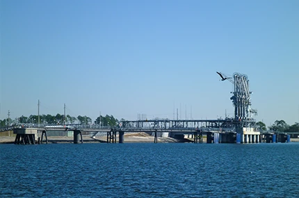 Industrial structure extends over water with piping and platforms; a bird flies overhead under a clear blue sky.