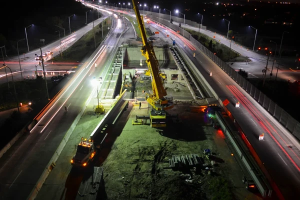 Nighttime construction scene showing a crane and workers on a highway overpass, with traffic on either side and bright work lights illuminating the area.