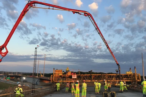 Construction workers in high-visibility clothing operate machinery and a concrete pump truck on a highway bridge under construction at sunset.