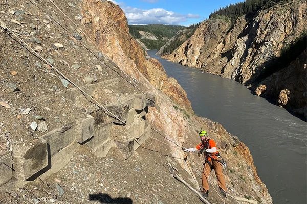 A worker in safety gear climbs a steep, rocky slope secured by ropes above a river running through a canyon.