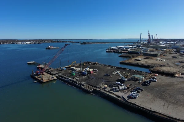 Aerial view of an industrial port with construction equipment, cranes, vehicles, and cargo along the waterfront under a clear blue sky.