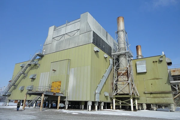 A large industrial building with metal siding, external staircases, and two tall rusted smokestacks, set on a snowy ground under a clear blue sky.