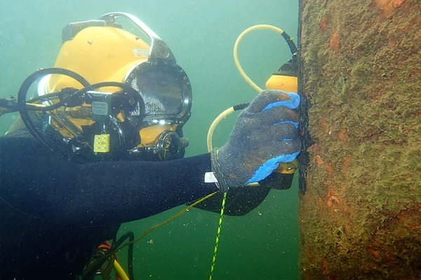 A diver in a helmet and gloves uses a handheld device to inspect or clean a large underwater pipe or structure covered with marine growth.