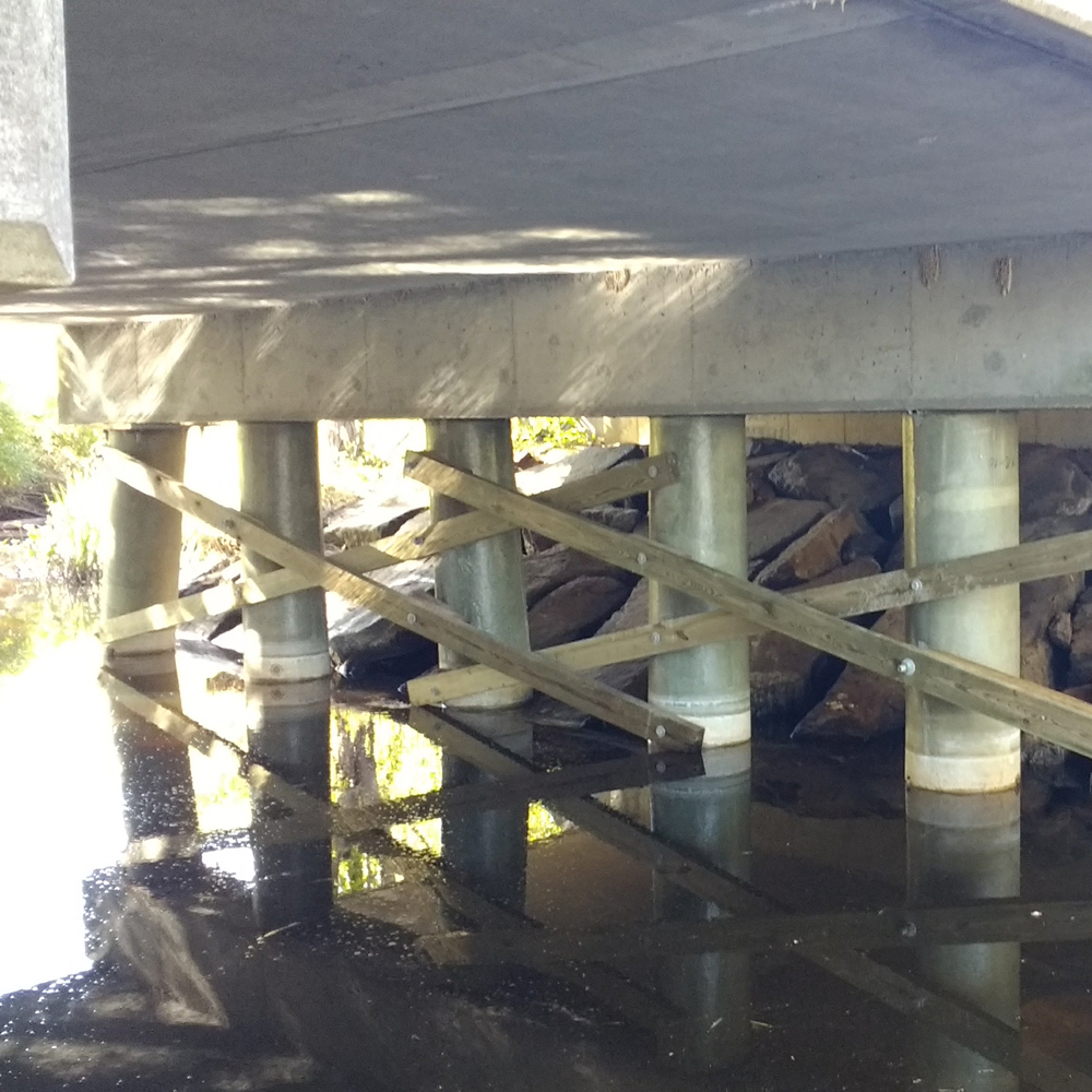 Concrete Brookfield bridge supported by round pillars and wooden cross-bracing, with rocks and water visible underneath the structure.