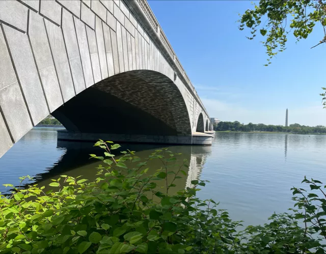 A stone arch bridge spans over a calm river, with green foliage in the foreground and a tall monument visible in the distant background.