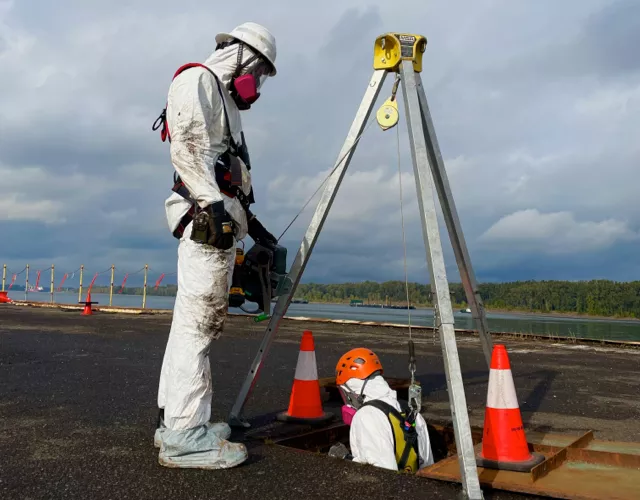 Two workers in protective suits and helmets operate near an open manhole, one entering while attached to a tripod hoist, with traffic cones marking the area.