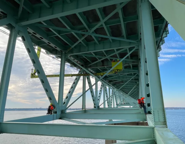 Two workers in safety gear conduct maintenance on the steel framework beneath a large bridge over water, with blue sky and clouds in the background.