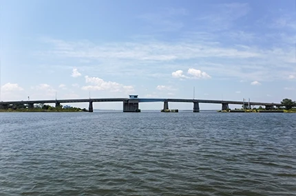 Concrete bridge spans a wide river under a partly cloudy sky, with water in the foreground and some trees visible on the shorelines.