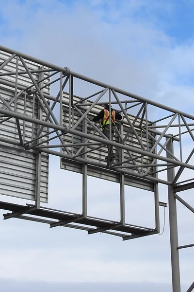 A worker wearing a safety vest is installing or repairing a large metal road sign structure high above the ground against a partly cloudy sky.