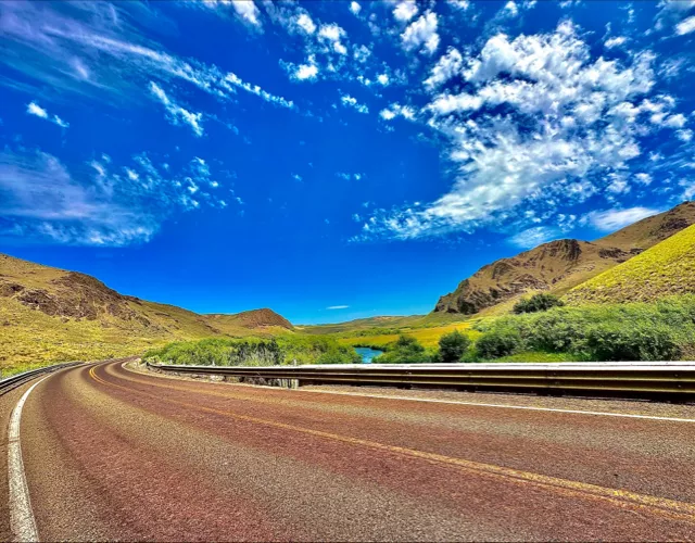 A winding road curves through a mountainous landscape under a bright blue sky with scattered clouds.