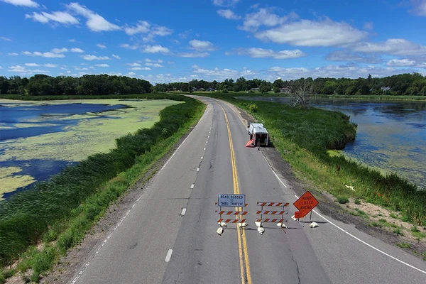 A rural road is closed with barricades and warning signs, surrounded by water and vegetation on both sides under a blue sky.