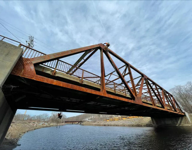 A rust-colored metal truss bridge spans over a calm river, with a partly cloudy sky above and some trees visible in the background.