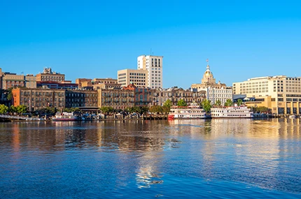 A city skyline with historic and modern buildings, a riverboat, and clear blue sky reflected in the calm water.