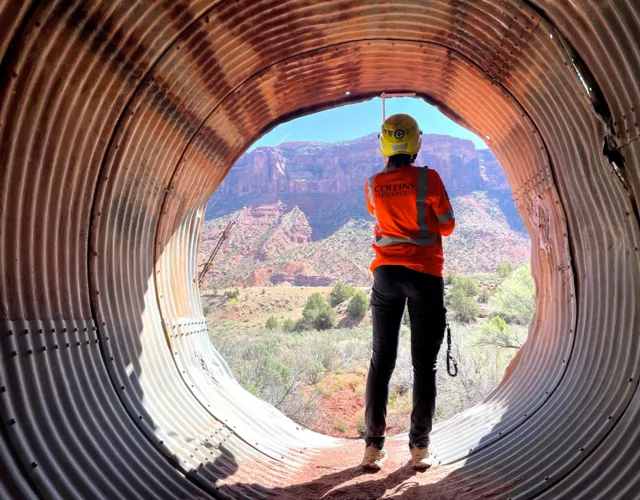 A person in safety gear stands at the end of a large corrugated metal pipe, facing a scenic canyon landscape with rocky cliffs and vegetation.