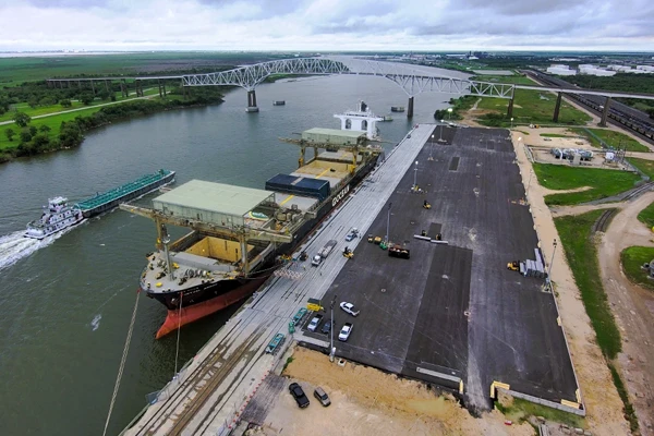 A cargo ship is docked at a port terminal, with a barge being pushed by a tugboat nearby; a large bridge spans the river in the background.