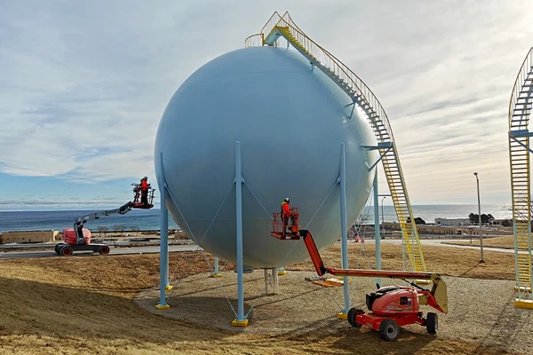Workers using boom lifts perform maintenance on a large spherical industrial storage tank supported by metal legs, with stairs and handrails attached.
