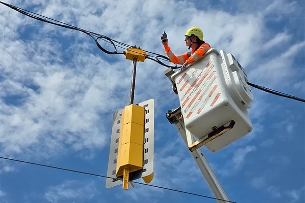 A worker in a safety helmet and vest operates from an elevated bucket truck, performing maintenance on overhead electrical wires against a blue sky with clouds.
