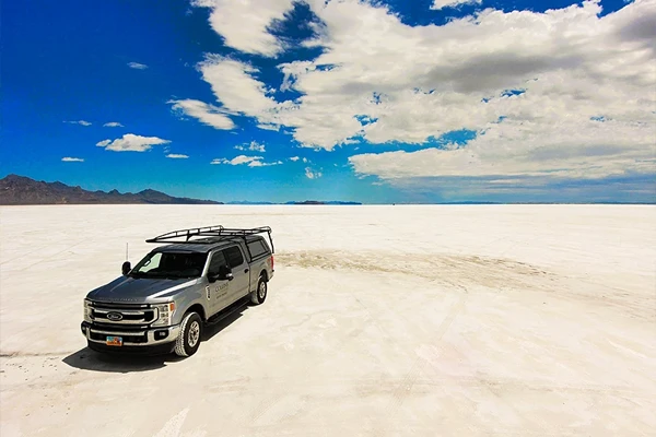 A gray pickup truck is parked on a vast, flat white salt plain under a partly cloudy sky with distant mountains on the horizon.