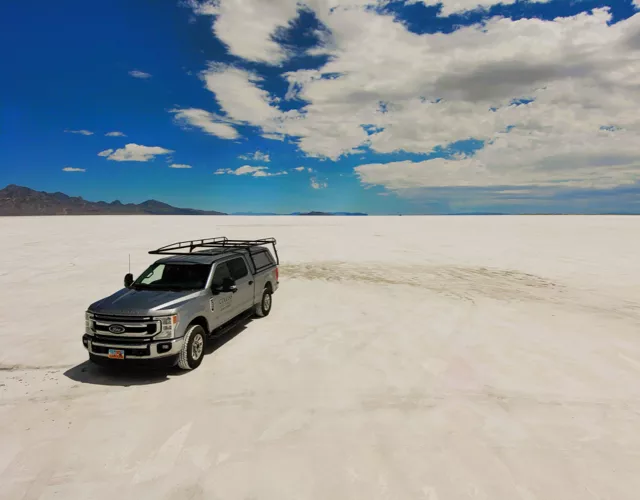 A silver SUV is parked on a vast, flat expanse of white salt with mountains and clouds in the background under a blue sky.