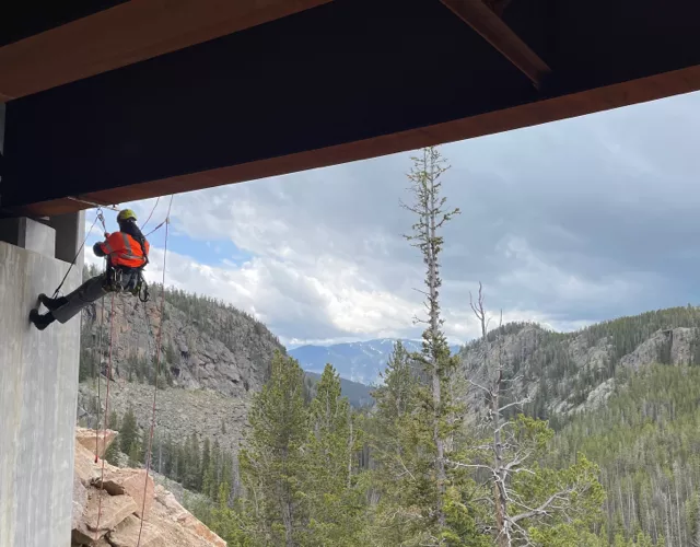 A person in safety gear rappels down a concrete structure under a bridge, with a forested mountain landscape in the background.
