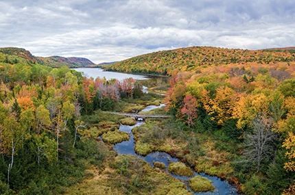 A river flows through a forest with autumn foliage, surrounded by rolling hills under a cloudy sky.