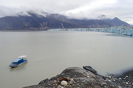 A small boat floats on a grey lake near a rocky shore, with a glacier and mountains in the background under a cloudy sky.