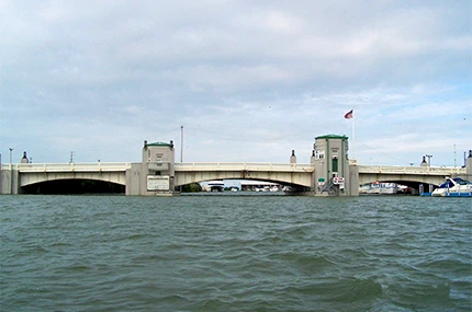 A drawbridge spans across a body of water under a cloudy sky, with visible bridge towers and a flag.