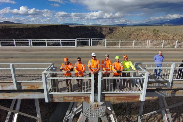 A group of seven people, most in orange safety gear, stand on a bridge with a canyon in the background under a partly cloudy sky.