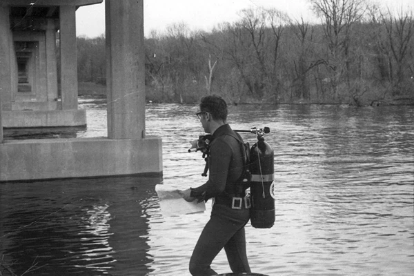 A person in scuba gear stands in shallow water near a bridge, holding a piece of paper, with trees visible in the background.