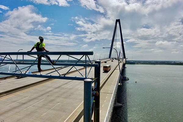 A worker in safety gear sits on a metal framework beside a large cable-stayed bridge over a body of water under a partly cloudy sky.