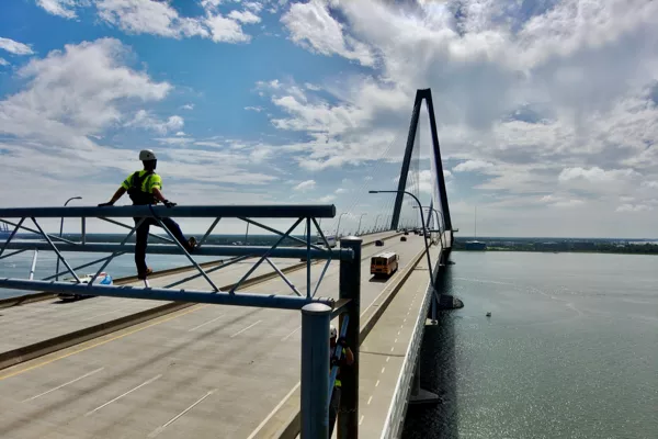 A worker in safety gear stands on a metal frame above a large bridge over water, with vehicles traveling on the bridge under a partly cloudy sky.