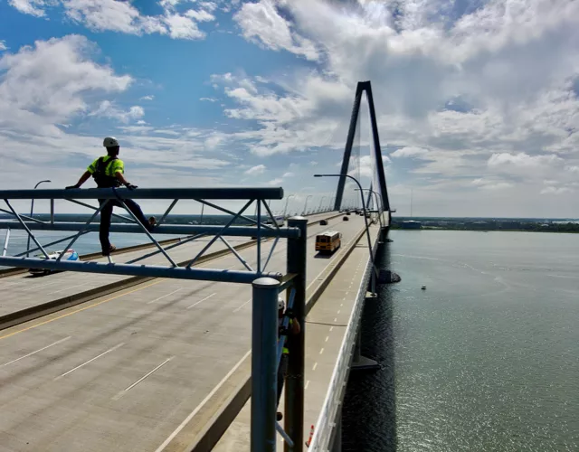 A worker in safety gear stands on a railing above a large bridge over water, with vehicles visible on the bridge under a partly cloudy sky.