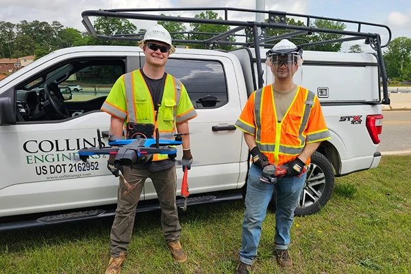 Two workers in safety gear stand in front of a Collins Engineering pickup truck; one holds a drone, the other holds a tool, with safety equipment visible.