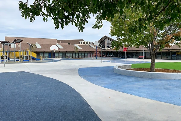 A school playground with a blue and white surface, basketball hoops, playground equipment, a tree, and a large building in the background.