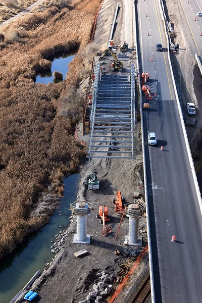 Aerial view of a highway under construction with heavy machinery, steel framework, and concrete columns next to a waterway and dry vegetation.