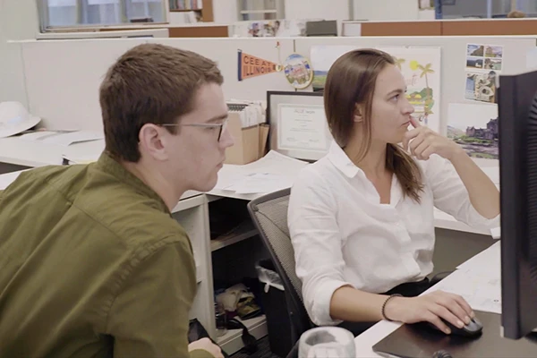 Two office workers sit at a desk, looking intently at a computer monitor in a modern open-plan office with cubicles.
