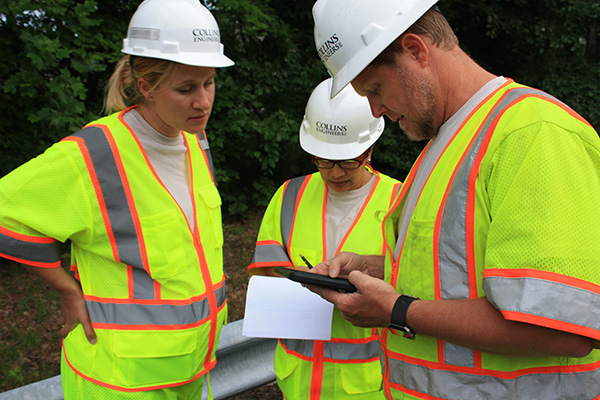 Three construction workers wearing safety vests and hard hats review information from a tablet featuring infrastructure asset management software and a notepad outdoors near greenery and a guardrail.