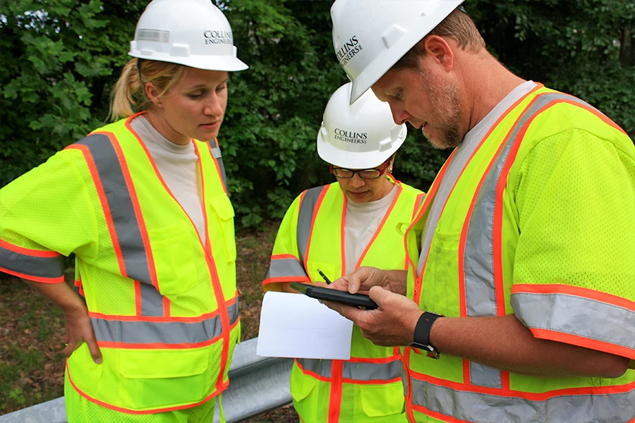 Three engineers wearing high-visibility vests and hard hats review documents and a tablet outdoors near greenery.
