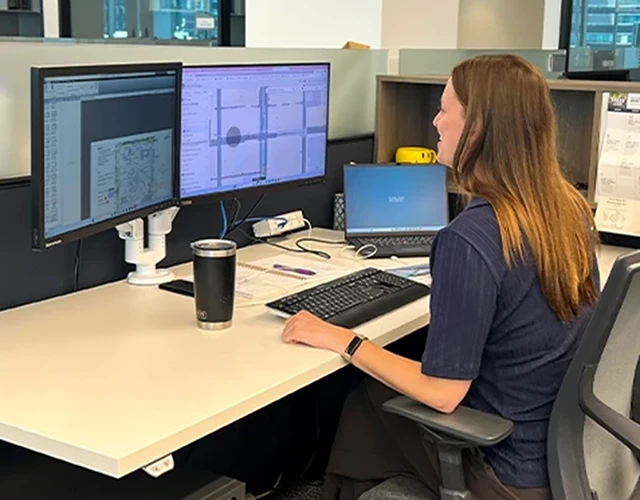 A woman sits at a desk working on dual monitors displaying maps and charts in a modern office setting.
