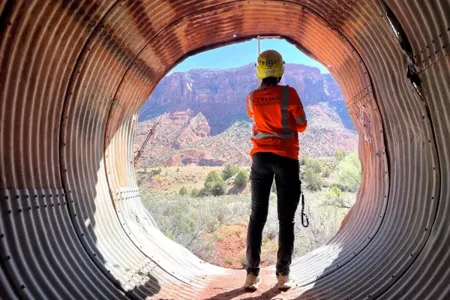 A person in safety gear stands inside a large corrugated metal pipe, looking out at a rocky landscape with cliffs and sparse vegetation.