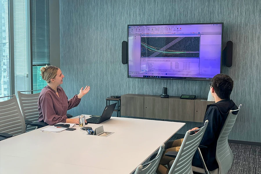 Two people sit in a conference room looking at a large screen displaying an engineering or architectural software program with diagrams.