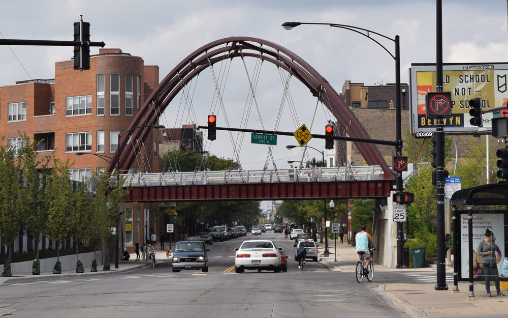 A city street with cars, cyclists, and pedestrians beneath an arched pedestrian bridge, surrounded by buildings and traffic lights.