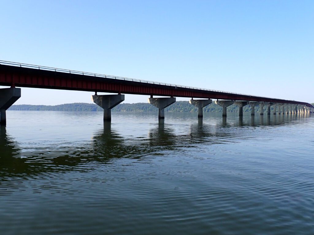 A long bridge with concrete supports across a wide body of water, with distant hills visible under a clear blue sky.